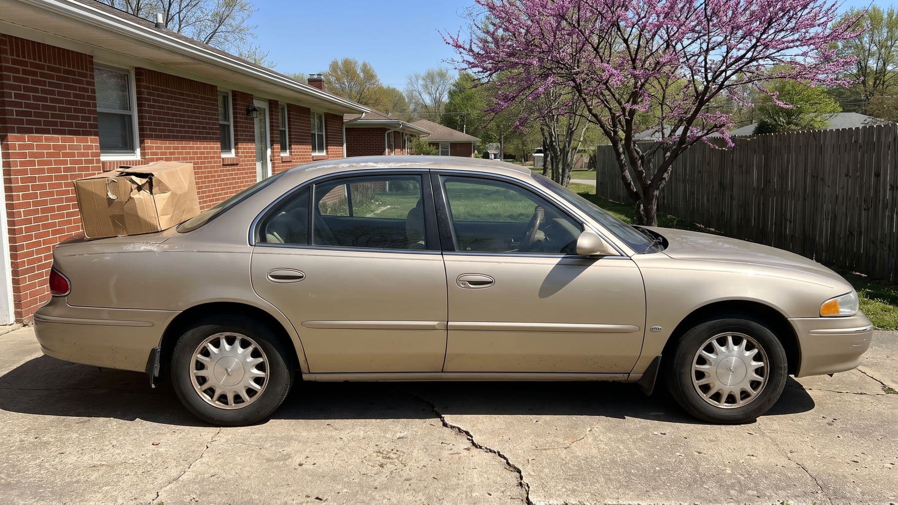 Illustration: a brown cardboard parts box resting on the trunk lid of a gold sedan in the driveway of a brick ranch house in Tulsa.