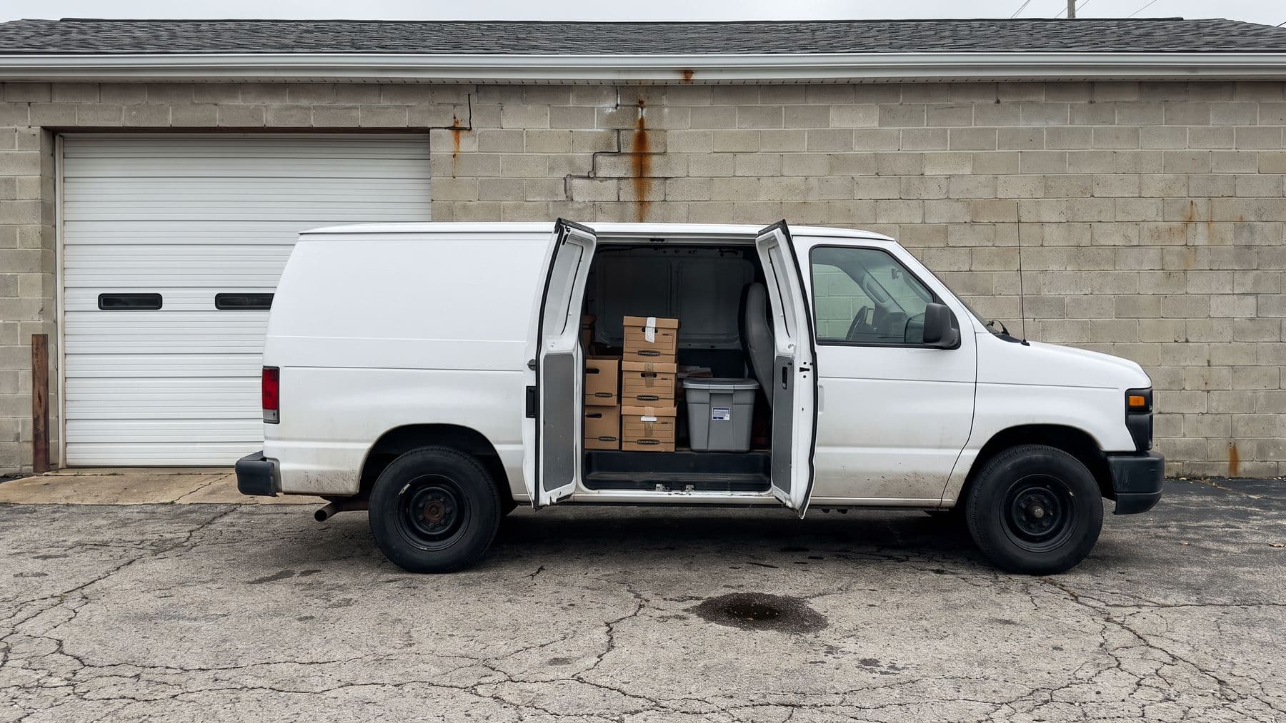 Illustration: a white cargo van with its side doors open on file boxes, parked on the cracked apron of a cinder-block repair shop in Indianapolis.
