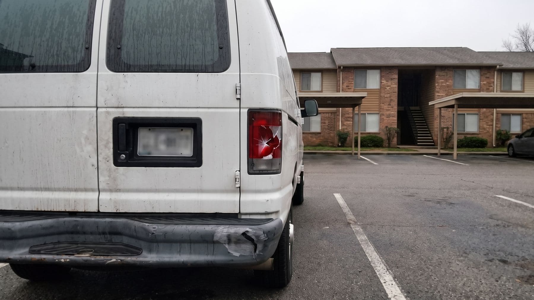 Illustration: the rear corner of a white cargo van with a scraped bumper and a cracked taillight in an apartment parking lot.