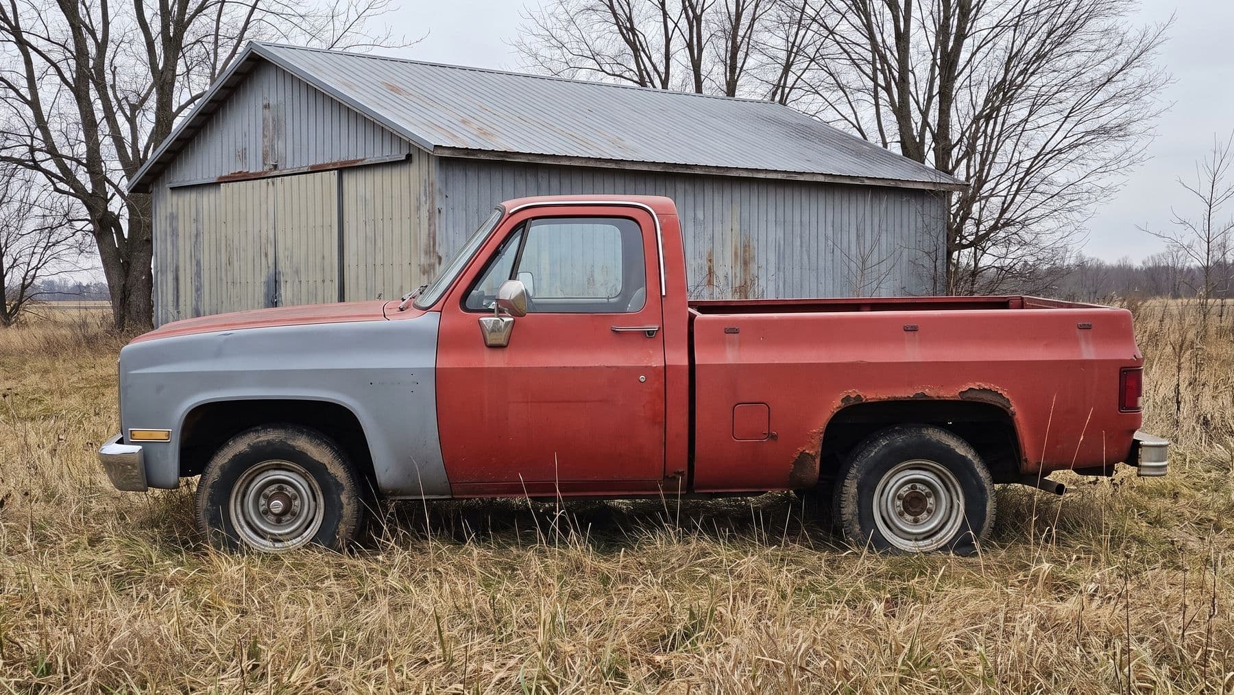 Illustration: a faded red 1980s pickup with two flat tires in the tall dead grass beside a pole barn in rural Michigan.